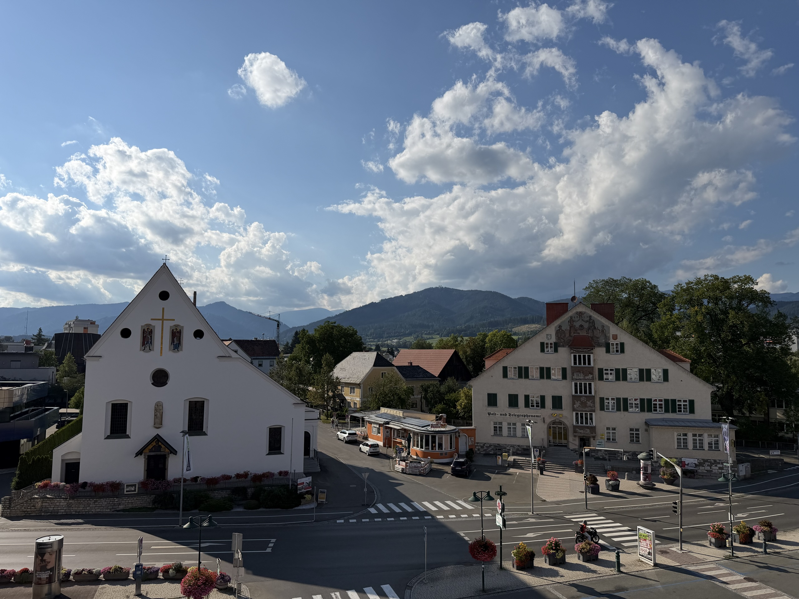 A church and neighboring building face a street with mountains beneath a cloudy sky.