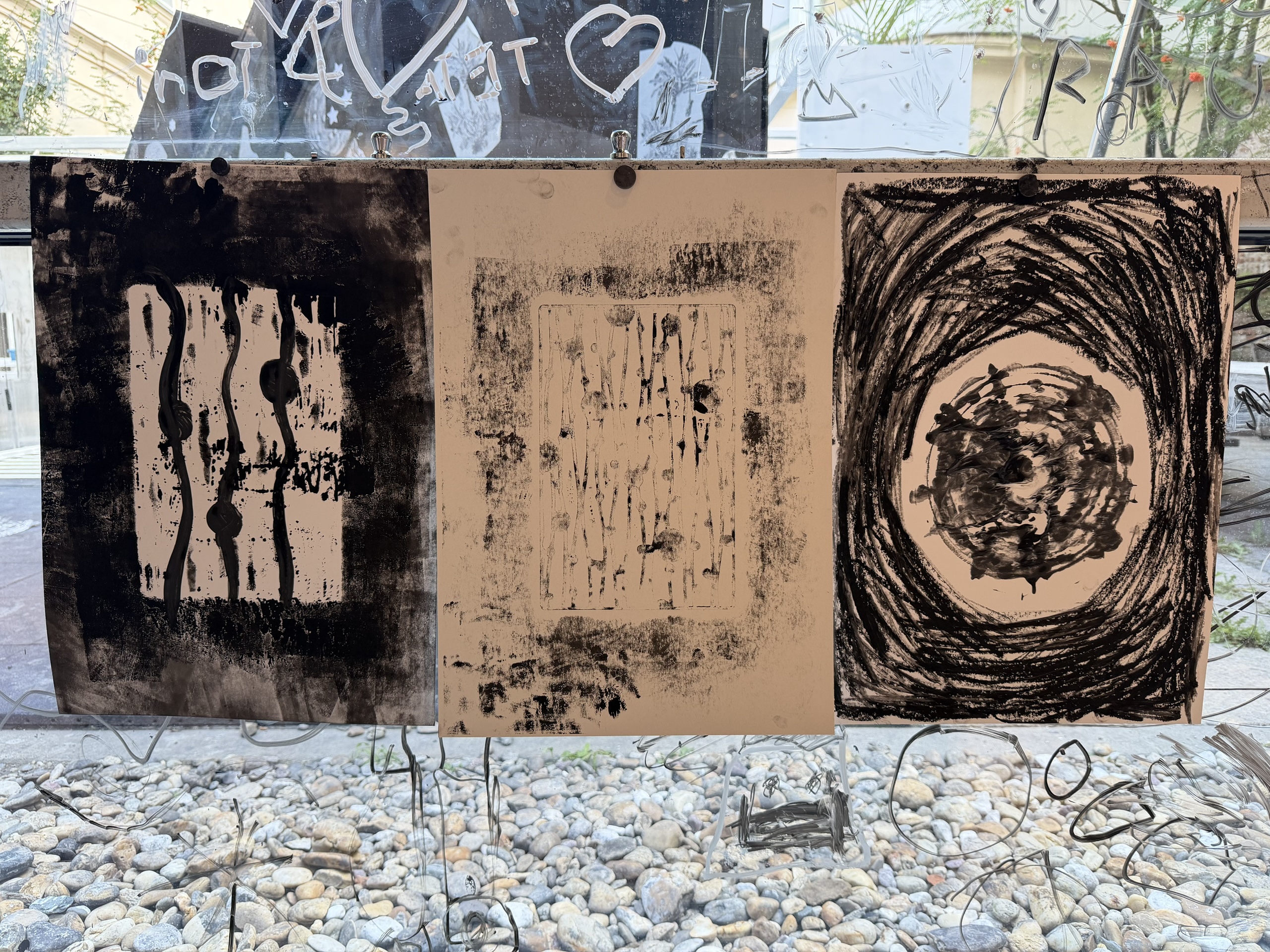 Three black ink prints hanging against a glass panel covered with drawings.