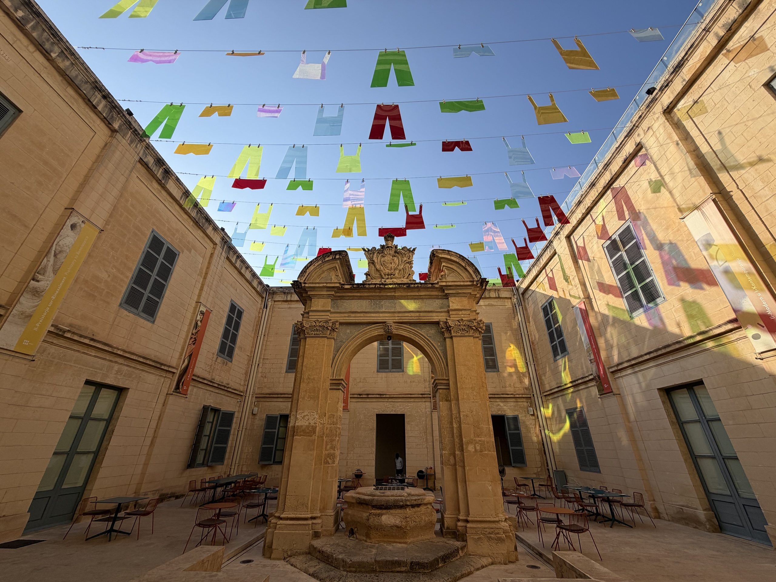 Colorful translucent clothing shapes hang above a stone courtyard and ornate arch.