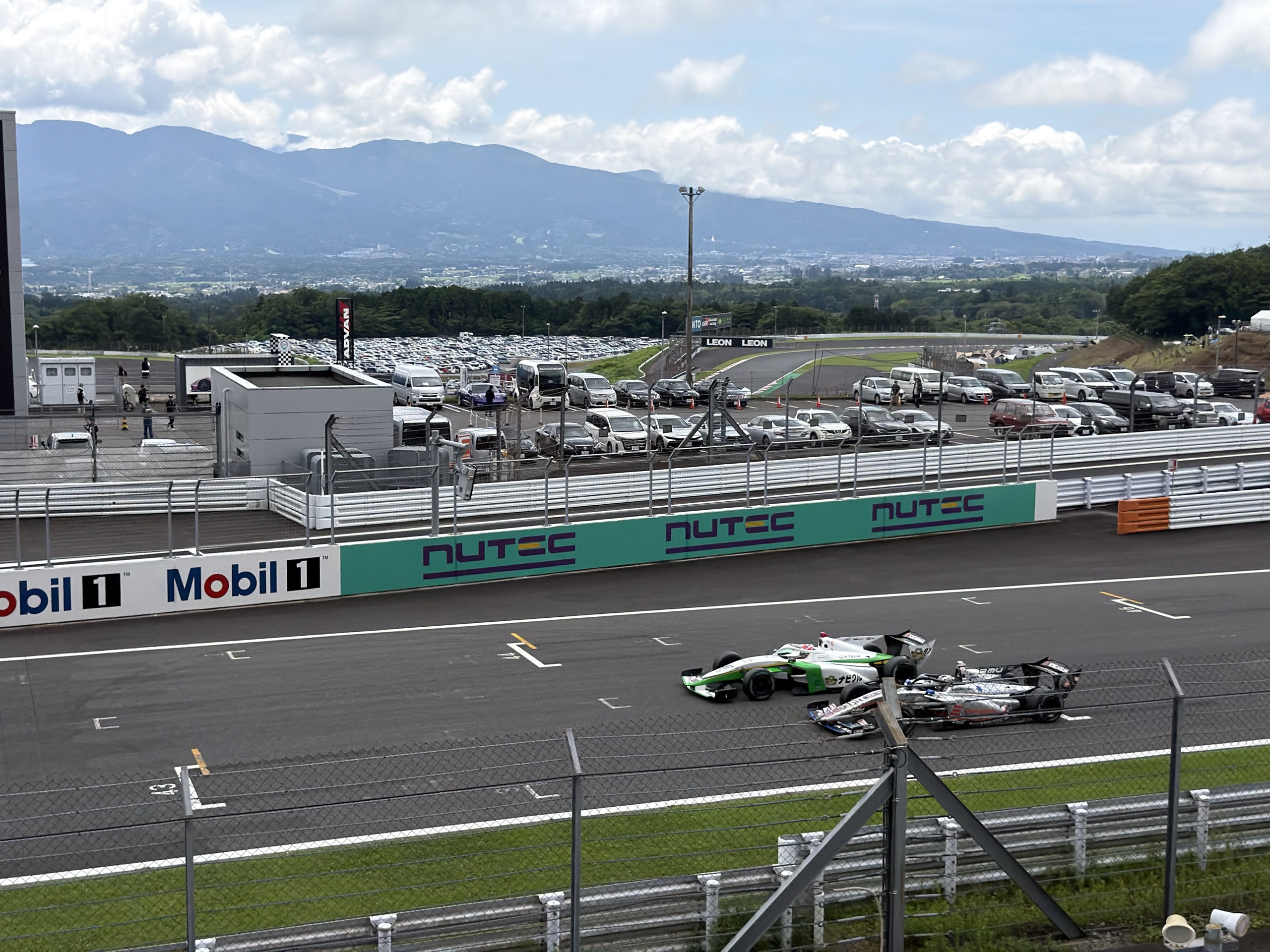 Two race cars on a racetrack, with parked cars and mountains beneath a cloudy sky.