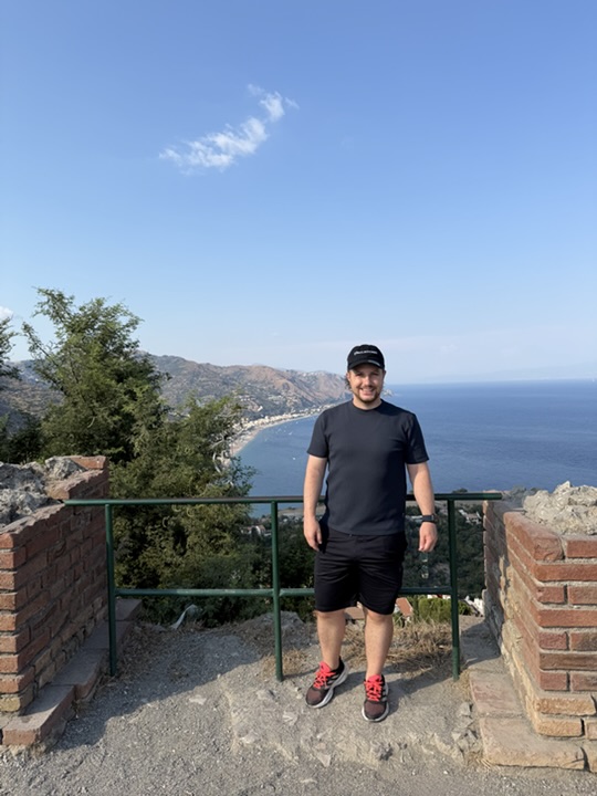 A man in a dark shirt and cap stands above a coastline under a blue sky.